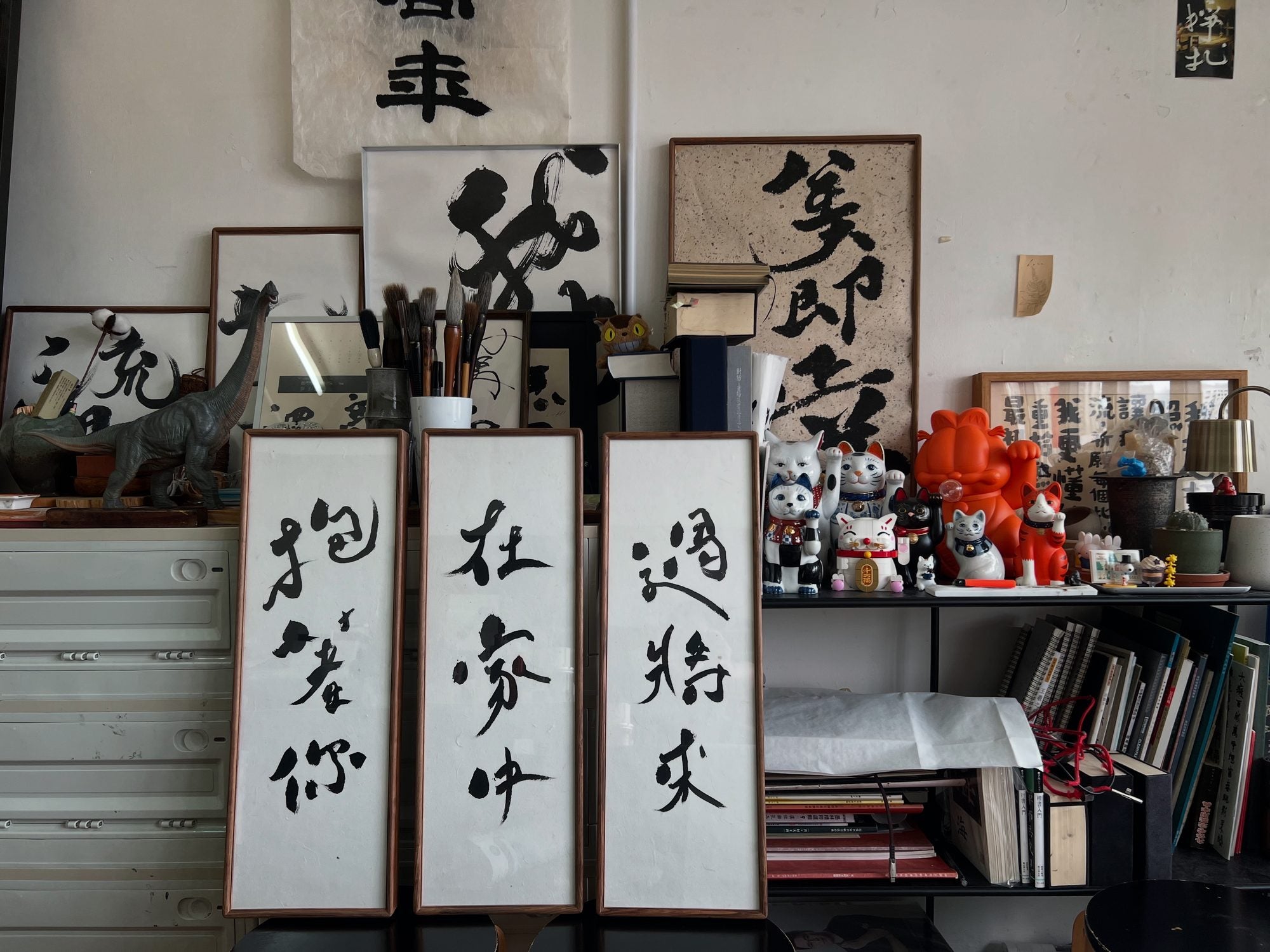 The Sellwordss studio: framed works stacked on shelves, jars of brushes, lucky-cat figurines, and a three-panel piece reading 抱緊你在家中過將來.