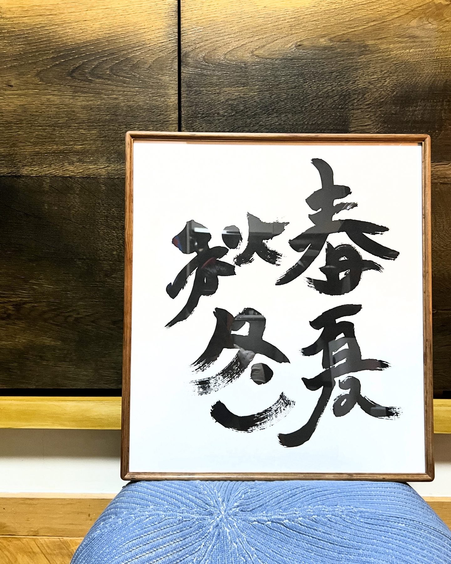 春夏秋冬 — Four Seasons. Four large grass-script characters in a wooden frame, resting on a blue knitted ottoman.