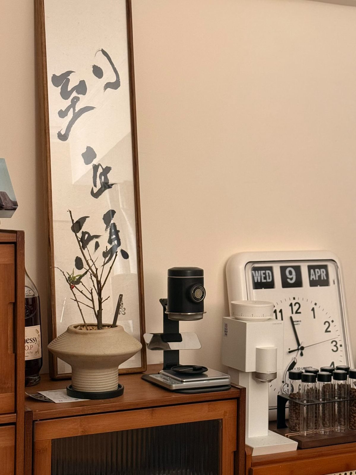 到白頭 — a tall narrow scroll standing on a wooden cabinet beside a coffee grinder, a flip clock and a small potted tree.