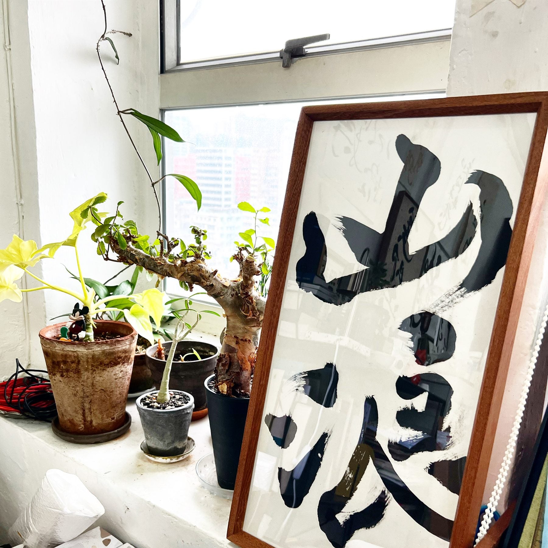 山浪 — Mountain Waves, a large framed piece leaning on a bright windowsill crowded with potted plants.