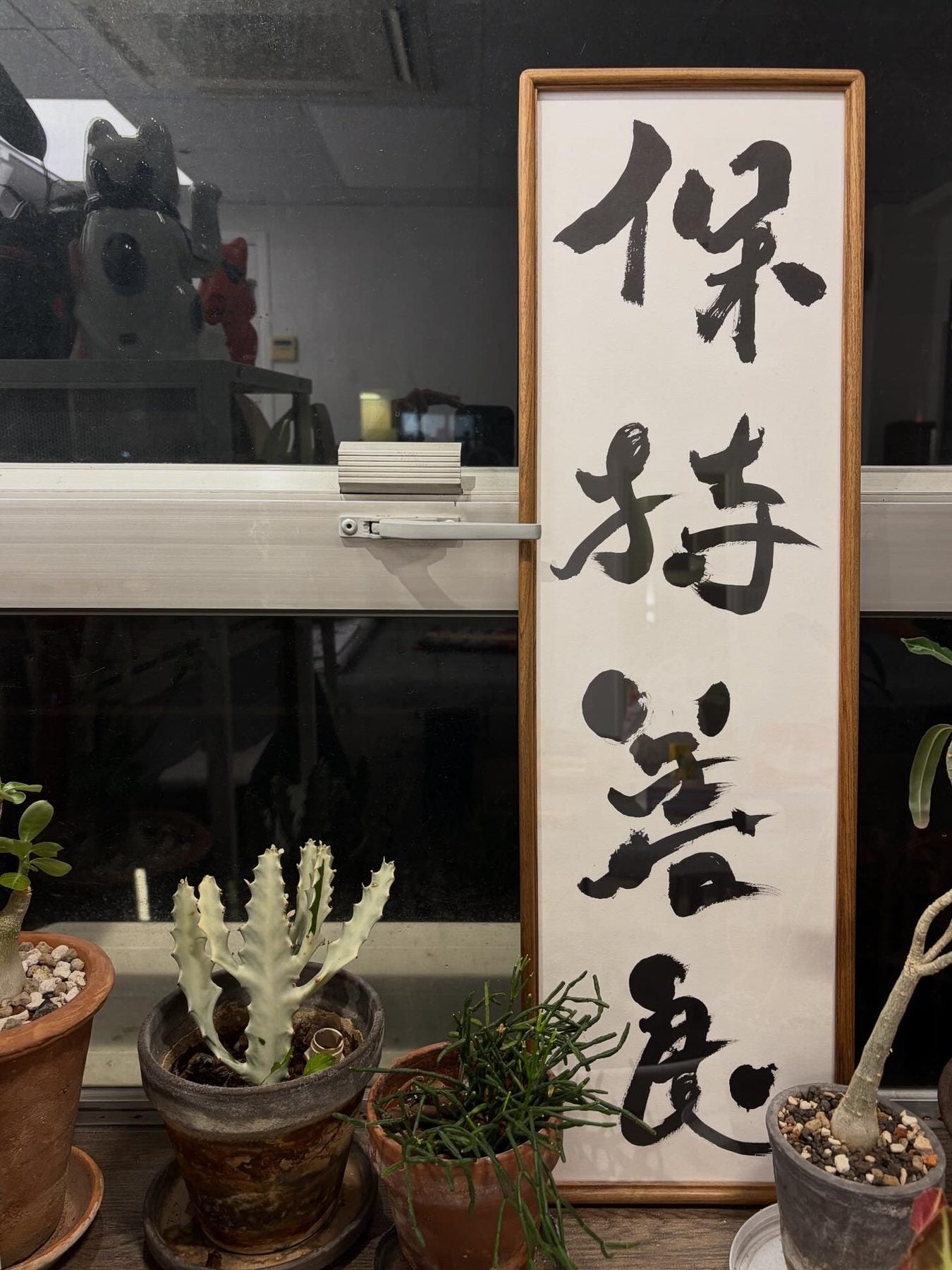 保持善良 — Stay Kind. A tall framed scroll of four characters, standing on a windowsill among potted plants.