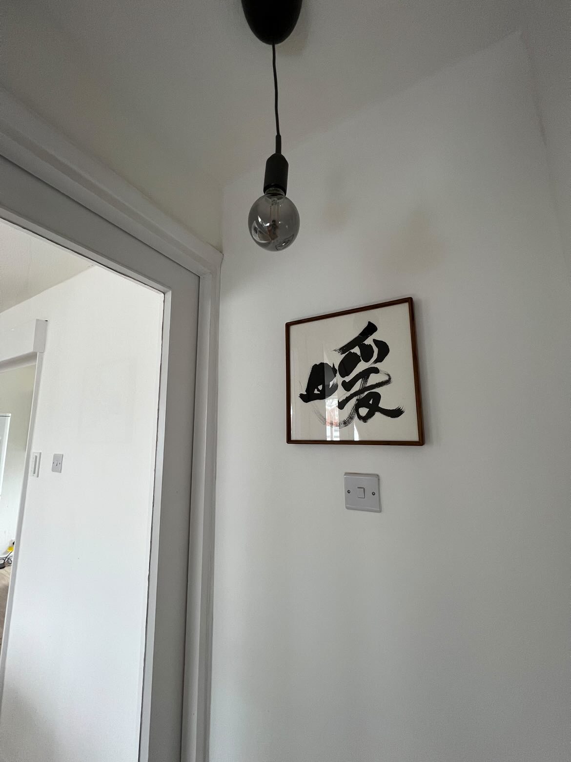 暖 — Warmth. A small framed character on a white hallway wall beneath a bare pendant bulb.